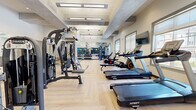 Located on Tulip Lane in Scotch Plains, NJ, this modern fitness center offers a comprehensive workout facility across a single, open floor. The space features a stylish herringbone-patterned light wood floor that runs throughout, contributing to a clean and contemporary aesthetic. The ceiling design incorporates exposed concrete beam accents contrasted against white surfaces, with recessed and linear pendant lighting ensuring the space is bright and well-illuminated.

The fitness center is thoughtfully organized to accommodate a wide range of exercise modalities. The cardio zone is lined with Spirit-brand treadmills, stationary bikes, and elliptical machines positioned along a wall of large windows, providing natural light and outdoor views of the surrounding community. Wall-mounted flat-screen televisions are positioned throughout for entertainment during workouts.

The strength training area features a multi-station cable machine system by Batca, adjustable benches, and a dedicated free weights section with a full rack of dumbbells and a variety of medicine balls stored on built-in shelving. Floor-to-ceiling mirrors span one wall, visually expanding the space and supporting proper form during exercise.

Overall, the fitness center presents a polished, fully appointed environment designed to support diverse fitness routines in a bright, welcoming atmosphere.