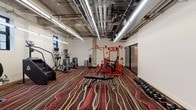 The fitness center at 121 Westmoreland Ave in White Plains, NY offers a well-equipped, single-floor workout space designed to accommodate a broad range of exercise needs. The interior is characterized by an industrial aesthetic, with exposed ceilings revealing ductwork, concrete structural elements, and long linear LED lighting that illuminates the space efficiently throughout. A bold, colorful striped carpet in deep reds, pinks, and earth tones covers the floor, adding a dynamic visual energy to the environment. Natural light filters in through a row of steel-framed windows along one wall, providing a connection to the outdoors while maintaining a focused workout atmosphere. The cardio section is generously stocked with treadmills, upright and recumbent stationary bikes, elliptical machines, a stair climber, and a stair stepper. Strength training is anchored by a prominent multi-station orange cable machine, complemented by an adjustable weight bench and a full rack of dumbbells arranged along the wall. A glass-partitioned area is visible adjacent to the main floor, suggesting a separate functional or stretching zone. An exercise mat and additional benches round out the available equipment, making this fitness center a comprehensive facility for both cardio conditioning and resistance training.