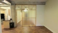 This single-floor space showcases a compelling blend of industrial and modern design elements. The most striking architectural feature is a floor-to-ceiling partition wall composed of frosted glass panels set within a warm wood frame, dividing the interior into distinct functional zones while maintaining an open, airy feel. The frosted glass allows diffused light to pass between areas, preserving a sense of spatial continuity throughout.

Exposed concrete beams run overhead, lending an industrial character that contrasts effectively with the smooth white walls and rich, dark hardwood-style flooring underfoot. The flooring extends consistently throughout the visible areas, creating a cohesive visual flow. A ceiling fan is mounted in the partitioned section, contributing to both comfort and casual ambiance.

A hallway is visible beyond the partition, hinting at additional rooms or storage areas accessible from the main space. The layout appears well-considered, balancing openness with defined zones. The overall ambiance is contemporary and understated, appealing to those who appreciate clean lines, raw architectural details, and versatile, flexible living or working environments.