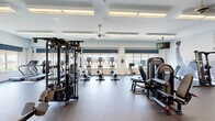 Located at 100 Fox Meadow Dr in Maple Shade, NJ, this fitness center offers a generously sized, single-floor workout facility designed to support a comprehensive exercise experience. The space is thoughtfully laid out to accommodate a wide variety of training styles, with clearly defined zones for cardio, strength, and functional training.

Large windows line the perimeter walls, flooding the interior with abundant natural light and providing open views of the surrounding outdoor landscape. Decorative roman shades with a polka-dot pattern add a touch of style to the bright, airy environment. Ceiling fans and recessed panel lighting contribute to a comfortable atmosphere throughout.

The cardio zone is well-stocked with treadmills, elliptical machines, stair climbers, and stationary cycling bikes, many equipped with individual display screens. The strength training area features a multi-station cable machine, a full rack of dumbbells, adjustable benches, and various selectorized weight machines. Rubber flooring throughout the space provides durability and safety underfoot.

Motivational wall art, including a large athletic mural and an inspirational Rocky Balboa quote, energizes the environment. Wall-mounted televisions are positioned throughout for added entertainment. The clean, modern design and thoughtfully arranged equipment make this fitness center a functional and inviting space for users of all fitness levels.
