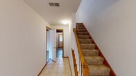 This three-floor home offers a practical and straightforward layout designed for comfortable everyday living. The main level features light tile flooring throughout the entryway and kitchen, with a staircase finished in warm wood tones and carpeted treads connecting all three floors. The kitchen is efficiently arranged in a galley style, featuring white cabinetry with wood trim, a double sink, tile flooring, and white appliances including a range and refrigerator, with a window providing natural light. A bathroom on the main level includes tile flooring, a vanity with white cabinetry, and a window with blinds. The upper floor is home to generously sized bedrooms, each featuring plush wall-to-wall carpeting, neutral painted walls, and windows that invite natural light. One bedroom includes a wide closet with bifold doors and a built-in hanging rod, offering ample storage. An open carpeted area adjacent to the staircase landing provides additional flexible space. The lower level presents a spacious, unfinished room with concrete flooring, painted cinder block walls, and a prominent structural wood beam overhead — well-suited for storage or future finishing. Throughout the home, warm wood baseboards and door trim provide consistent, cohesive detailing.