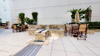 This sophisticated outdoor courtyard space offers a serene retreat within a modern urban setting. The grounds are thoughtfully designed with a combination of elegant hardscaping and lush landscaping, creating a welcoming atmosphere for relaxation and social gathering.

At the heart of the space sits a striking stacked-stone water feature with a cascading waterfall element, flanked by built-in seating ledges and accented with decorative glass rock at its base. Adjacent to the water feature, a built-in fire pit adds warmth and ambiance, making the space enjoyable across seasons. Bar-height seating with patio umbrellas is conveniently positioned nearby, providing a comfortable spot for outdoor dining or conversation.

The patio is finished with large-format stone tile flooring and complemented by raised stone planters housing a variety of manicured shrubs, seasonal flowers, and mature trees. Oversized concrete planters add vertical greenery throughout. Brick detailing along the base of the surrounding structures adds a classic architectural touch, while a glass-door entry leads to a tiled interior corridor. The courtyard is enveloped by a multi-story building complex, offering a sense of privacy while maintaining an open, airy feel under natural daylight.