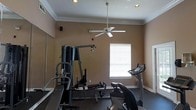 This dedicated fitness space is designed to support a well-rounded workout routine within a single-floor layout. The room features warm, tan-toned walls accented by white crown molding and clean white baseboards, lending a polished and tidy appearance. Recessed lighting is installed in the ceiling alongside a centrally mounted ceiling fan, ensuring the space remains well-lit and comfortable during exercise sessions. A large wall mirror spans one side of the room, creating a sense of expanded space while allowing users to monitor their form. Natural light filters into the room through a window fitted with white horizontal blinds, adding brightness without harsh glare. The fitness equipment includes a multi-function cable and weight machine, ideal for a variety of strength training exercises, as well as cardio equipment such as a treadmill and a stationary bike, offering options for both endurance and resistance training. The flooring appears to incorporate a darker surface material suited for high-activity use. A small speaker is mounted on the wall, supporting an audio setup for an enhanced workout atmosphere. Overall, the space is thoughtfully arranged to maximize functionality.