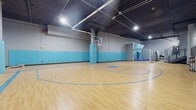 This indoor futsal facility offers a generously sized, single-level playing surface covered in green artificial turf, complete with clearly marked white boundary lines, a center circle, and goal boxes at each end. Two full-size portable goals are positioned at opposing ends of the pitch, making the space ready for competitive or casual play. The structural aesthetic is defined by exposed concrete walls and ceiling, lending an industrial character that is both raw and functional. Overhead, a grid of recessed and surface-mounted fluorescent panel lights ensures even, bright illumination across the entire playing area. One wall is dramatically transformed by a large-scale graphic mural featuring bold blue and white geometric shapes — hexagons and sharp angular forms — alongside the energetic phrase "Live, Work, Play" rendered in dynamic lettering. This eye-catching artwork adds a vibrant, motivational atmosphere to the space. Along one side wall, player benches and a utility rack provide practical amenities for teams and participants. The space is well-suited for organized matches, training sessions, or recreational leagues, combining athletic functionality with a visually engaging environment that encourages active use.