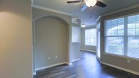 This single-floor residence showcases a thoughtfully designed interior that balances style and functionality. The living area immediately draws attention with its elegant arched architectural detail, which serves as a refined passageway connecting spaces and adding a timeless character to the layout. Crown molding runs along the ceilings throughout, lending a polished, upscale finish to each room.

Dark hardwood flooring extends across the open-plan space, providing a rich contrast against the neutral warm-gray walls and crisp white baseboards and trim. A ceiling fan with a light fixture is centrally installed, offering both practical airflow and ambient lighting.

Large windows dressed with horizontal blinds line one side of the space, allowing generous natural light to flood the interior and creating a bright, airy atmosphere. The open layout transitions smoothly into an adjoining area, partially visible beyond a half-wall or breakfast bar, hinting at a connected kitchen or dining zone. A second ceiling light fixture is visible in this adjacent space, reinforcing the continuity of the design.

Overall, the space presents a clean, well-maintained interior with consistent architectural detailing, warm tones, and a functional open-concept flow that makes it feel welcoming and spacious.