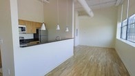 This single-floor space showcases an open-plan design that seamlessly connects the kitchen and living areas. The kitchen features a dark granite breakfast bar with a sleek faucet, natural wood cabinetry, and stainless steel appliances, including a full-sized refrigerator. Three white conical pendant lights hang above the bar, providing focused task lighting while adding a contemporary aesthetic touch.

The living area extends beyond the kitchen partition, offering a generous, unobstructed space with light-toned hardwood flooring that runs throughout, lending warmth and continuity to the interior. The ceilings are notably high, contributing to an airy, expansive atmosphere, and are distinguished by an exposed cylindrical ventilation duct — a hallmark of industrial loft-style architecture. The walls are finished in a clean, neutral tone, providing a versatile backdrop. An electrical panel is discretely set into the wall, and recessed infrastructure details reflect thoughtful, functional design. Overall, the space balances modern finishes with industrial character, creating a bright and open environment.