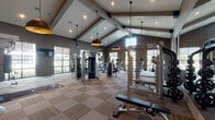 This fitness center in Cypress, TX, is a spacious, single-floor facility designed to accommodate a wide range of workout preferences. The architectural highlight is the dramatic vaulted ceiling supported by exposed wood beams, which lends the space a grand, open feel. Recessed lighting and stylish gold-interior dome pendant lights illuminate the facility, complementing the warm, neutral-toned walls painted in deep gray with bold geometric accent patterns in select areas.

The flooring throughout consists of modular carpet tiles in a checkered tan and brown pattern, providing both comfort and durability underfoot. Full-length mirrors along one wall visually expand the space and allow users to monitor their form during workouts.

The cardio zone features a row of modern treadmills, ellipticals, and stair climbers, each equipped with individual screens, all positioned near large windows that invite natural light and exterior views. The strength training area is outfitted with cable machines, a power rack, and weight benches. A dedicated functional training nook includes a wall-mounted rig with kettlebells, medicine balls, resistance bands, and a BOSU ball.

Entry is through sleek glass-paneled double doors that connect to an adjacent bright, wood-floored corridor. The overall ambiance is polished, modern, and energizing.