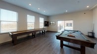 This single-floor amenity space is designed with both function and leisure in mind, offering residents a well-appointed environment for entertaining and recreation. The kitchenette area is outfitted with rich dark cabinetry, light granite countertops, a stainless steel double-basin sink, a full-size refrigerator, and a dishwasher. A large statement clock adorns the wall, adding an industrial-chic character to the space. A generous island with a curved granite top and rustic bar stools provides a comfortable gathering spot for socializing. The game room is expansive and open, anchored by a wood-framed pool table with cues and a rack, as well as a full-length shuffleboard table. A wall-mounted flat-screen TV is positioned for easy viewing from multiple vantage points. Natural light flows into the game room through multiple windows with blinds and a sliding glass door that opens to an outdoor balcony or terrace, extending the social space beyond the interior. Throughout, durable wood-look flooring ties the areas together with a warm, contemporary aesthetic. Recessed lighting and clean white walls keep the atmosphere bright and inviting, making this a versatile space well-suited for casual hangouts and community events alike.