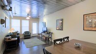 This unique single-floor space makes an immediate impression with its standout corrugated metal ceiling, which lends an industrial yet warm aesthetic throughout the open-plan interior. Dark hardwood floors run continuously underfoot, grounding the space and providing a rich contrast to the light-toned walls.

The open layout seamlessly connects the dining and living areas, creating a cohesive flow ideal for both everyday living and entertaining. The dining area features a solid wood table with ample seating, positioned to take full advantage of the open sightlines across the space. The living area is anchored by a comfortable seating arrangement set atop a soft area rug, with a media setup nearby for relaxed viewing.

Natural light enters generously through multiple windows fitted with white blinds, brightening the interior while a ceiling fan with a light fixture ensures comfortable airflow. Track lighting along the ceiling provides additional illumination and highlights the architectural character of the metal overhead detail.

Wall art adds personality and a creative touch throughout, complementing the space's eclectic, loft-inspired character. Overall, the space offers a well-balanced blend of industrial design elements and comfortable, functional living — making it a visually engaging and practical environment.