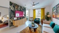This single-floor residence showcases a vibrant, eclectic design aesthetic that seamlessly blends bold artistic choices with modern finishes throughout. The open-concept kitchen is a standout, featuring a two-tone cabinetry scheme with dark lower cabinets and bright white uppers, a patterned blue tile backsplash, stainless steel appliances, a large center island with a white quartz countertop, and sleek pendant lighting — making it both a functional and visually striking space. The adjacent living area is filled with natural light from large windows dressed in rich gold curtains, and is anchored by a dramatic tropical botanical accent wall that frames the entertainment area. The room carries an artistic, gallery-like energy, with bold wall art and lively color accents throughout. The bedroom continues the expressive design with a delicate floral wallpaper accent wall, warm gold and neutral tones, a ceiling fan, and a window that brings in soft natural light and outdoor views. The bathroom is clean and contemporary, finished with light tile flooring, white cabinetry with matte black hardware, a framed mirror, and a shower-tub combination. Light wood-look flooring flows consistently throughout the space, tying the design together cohesively.
