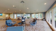 Located at 2350 Cypress Pond Rd in Palm Harbor, Florida, this well-appointed single-floor clubhouse offers a generous and thoughtfully designed interior space. The layout flows seamlessly from a welcoming lounge area to a professional reception zone, creating a versatile environment suited for both relaxation and community engagement.

The main living area is anchored by a patterned carpet and furnished with sleek, modern seating in blue and charcoal tones, accented by chrome-framed chairs arranged around low tables. An expansive bank of floor-to-ceiling windows lines the perimeter, flooding the space with natural light and offering unobstructed views of lush greenery and manicured landscaping outside.

The reception area features soaring ceilings with a dramatic abstract mural adorning a full-height accent wall, paired with a marble-topped counter and dark wood cabinetry. Recessed lighting throughout provides consistent, warm illumination across all areas.

A separate dining alcove with round tables and upholstered chairs opens directly to an outdoor pool area through glass-paneled doors, blurring the line between indoor comfort and Florida's natural outdoor environment. Soft teal accent walls complement the neutral flooring and bright white trim, contributing to the space's clean, contemporary aesthetic.