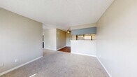This single-floor, one-bedroom, one-bathroom unit offers a practical and efficiently organized layout. The living room features neutral-toned carpet, textured ceilings, large windows with horizontal blinds that allow natural light to filter in, and a wall-mounted HVAC unit. The space opens toward the kitchen area, which is separated by a half-wall breakfast bar with a light-toned countertop. The kitchen is equipped with blue-gray cabinetry, a white electric coil range, a full-size refrigerator, a single-basin sink, and a range hood overhead. Hardwood-style flooring runs through the kitchen and connects to the dining area, which features a ceiling fan with light fixture. The bedroom is a comfortable size, carpeted, and includes a window that brings in natural light. A dedicated closet provides storage with dual hanging rods across two sections and hardwood-style flooring. The full bathroom includes a single-sink vanity with gray cabinetry, a toilet, and a full tub-shower combination with a white surround, complemented by hardwood-style flooring throughout.