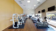 This fitness center offers a generous, open layout designed to accommodate a full range of exercise equipment and workout styles. The space features hardwood-style flooring throughout, providing a clean and durable surface that complements the room's bright, welcoming atmosphere. Elevated ceilings with recessed panel lighting contribute to an airy, open feel, while a bold yellow accent wall adds a energizing pop of color against the otherwise neutral white walls.

Along one side of the room, a selection of white cable and weight machines provides options for targeted strength training, each positioned on protective rubber mats. The opposite side is dedicated to cardio equipment, including treadmills and elliptical machines, arranged to take advantage of natural light streaming in through windows that offer views to the outdoors. A wall-mounted television is positioned for convenient viewing during cardio sessions.

A large mirror along the back wall visually expands the space and supports functional training, while a rack of dumbbells and exercise balls are visible in the background. The overall ambiance is clean, functional, and well-organized, making it a practical environment for a variety of fitness routines.
