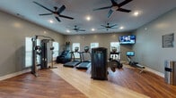This single-floor fitness center is thoughtfully divided into multiple functional workout zones, offering a comprehensive exercise experience. The main cardio area features a lineup of premium True-brand treadmills, stair climbers, ellipticals, and rowing machines, all set atop warm wood-look flooring. Multiple large ceiling fans and recessed lighting keep the space bright and well-ventilated, while wall-mounted flat-screen TVs provide entertainment during workouts. Natural light flows in through several windows fitted with white blinds, enhancing the open and airy atmosphere. A dedicated free weights area includes a full dumbbell rack and adjustable bench, alongside a plyo box and cable machine for functional strength training. A separate cycling studio houses a row of Cascade spin bikes, its own ceiling fan, and a wall-mounted TV, finished with durable dark concrete-style flooring. An additional multi-purpose studio space features large wall mirrors, a full assortment of stability balls, resistance bands, medicine balls, and a speed bag station, making it ideal for group fitness or individual cross-training sessions. Frosted glass French doors connect the distinct zones, contributing to a polished, modern aesthetic throughout the facility.