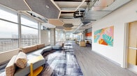 The Rise Community Lounge with Kitchen is a generously sized, single-floor amenity space located in Seattle, WA, designed to foster community gathering and social interaction. The open-plan layout is anchored by a striking industrial-modern aesthetic, featuring exposed concrete columns, visible ductwork, and soaring ceilings that lend an airy, loft-like character to the space.

Distinctive hexagonal pendant light fixtures with integrated LED lighting are suspended throughout, complementing the bold, patterned carpet that grounds the various activity zones below. Large floor-to-ceiling windows wrap around the perimeter, flooding the interior with abundant natural light and offering sweeping panoramic views of the Seattle skyline and surrounding cityscape.

The lounge area is furnished with modular sectional sofas and accent seating, creating flexible social zones suited for casual gatherings or relaxed conversation. A fully equipped kitchen island with bar seating, warm wood cabinetry, and stainless steel appliances anchors the culinary area, making the space ideal for events and entertaining.

Additional highlights include a dedicated dining and work area with tables and chairs positioned along the windowed walls, a large-format display screen mounted on a light wood feature wall with bold orange accent lines, and vibrant botanical wall art that adds a lively, colorful focal point. Glass doors open directly to an outdoor terrace with additional city views.
