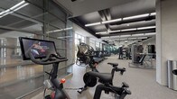 Located at 951 Fell St in Baltimore's vibrant Harbor East neighborhood, this contemporary fitness center offers a well-appointed, single-floor layout designed to support a wide range of workout routines. The space features a striking industrial-modern aesthetic, with a dark exposed ceiling accented by linear LED lighting that illuminates the floor throughout. Neutral-toned carpet tiles cover the flooring, contributing to a clean and professional environment.

The cardio zone is lined with an impressive row of treadmills, ellipticals, StairMaster climbers, and interactive cycling bikes equipped with screen displays for on-demand classes. Floor-to-ceiling glass partitions divide sections of the facility, adding visual openness while defining distinct workout areas. Large windows along the perimeter allow natural light to fill the space and provide views to the outdoors.

The strength training area includes a Smith machine, cable stations, Precor equipment, TRX suspension systems, a wall-mounted stall bar, and a well-stocked dumbbell rack ranging across multiple weights. Rowing machines, stability balls, medicine balls, resistance bands, and yoga mats round out the functional fitness offerings. Full-length mirrors are strategically placed throughout, enhancing the sense of space and supporting proper form. Changing room access is also available within the facility.