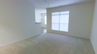 This single-story space features an open-plan layout that seamlessly connects the main living area to an adjacent dining space. The living area is finished with neutral-toned wall-to-wall carpeting, contributing to a clean and understated aesthetic. A large, multi-pane window dominates one wall, allowing an abundance of natural light to fill the room, enhancing the sense of openness throughout.

A half-wall partition separates the living space from the dining area, which features hard flooring and is illuminated by an elegant chandelier suspended from the ceiling. This subtle division maintains visual connectivity between the two areas while providing a clear sense of defined zones. A ceiling fan fixture is also present in the living area, adding both functional airflow and a finishing detail to the ceiling.

The overall color palette is neutral, with soft beige tones on both the walls and flooring, creating a calm and versatile environment. The single-floor layout promotes easy accessibility and flow. The abundant natural light combined with the open design gives the space an airy, welcoming ambiance suited to a variety of interior configurations.