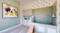 Located at 1830 Homerun Rd in Kissimmee, FL, this purpose-built pet spa is a charming and fully equipped grooming space designed with both functionality and style in mind. The room is anchored by a raised dog wash tub clad in sage-green square tiles, complete with a flexible hose attachment and a convenient wooden step stool for easy pet access. Adjacent to the tub, light wood cabinetry with matte black hardware provides ample storage above and below a white quartz countertop, which houses a matte black sink and wall-mounted pet dryer — keeping all grooming essentials within reach. The flooring features a distinctive hexagonal tile pattern in soft grey tones, adding visual interest underfoot. Whimsical hand-drawn dog-print wallpaper adorns the upper walls, lending the space a playful and welcoming personality, while sage-green painted lower walls provide a cohesive, calming palette. A large floor-to-ceiling window and glass door flood the room with natural light and offer sweeping views of the beautifully landscaped outdoor grounds below. A bold, oversized canine-themed artwork adds an artistic focal point. A wall-mounted mini-split air conditioning unit ensures year-round comfort, making this pet spa as practical as it is delightful.