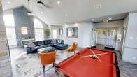 This single-floor recreational lounge is thoughtfully designed to serve as a premier social and entertainment hub. The space is anchored by a full-size pool table with a bold red felt surface, immediately drawing attention upon entry and setting an energetic, welcoming tone. Adjacent to the game area is a generously sized seating lounge, furnished with a large navy blue leather sectional sofa, complementary cognac leather accent chairs, and a tufted coffee table, all arranged atop a softly patterned area rug. The vaulted ceilings, accented by a ceiling fan, lend an open and airy quality to the room, while a striking arched window along with multiple additional windows floods the space with natural light. Recessed lighting throughout ensures the room remains well-lit during evening use. The walls are finished in a clean, neutral gray palette that complements the eclectic furnishings. A decorative framed mirror and tasteful greenery add refined touches to the overall aesthetic. French-style glass doors lead to adjoining areas, enhancing connectivity within the space. The combination of game amenities, comfortable lounge seating, and thoughtful architectural details makes this an inviting and versatile gathering space.