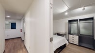 This thoughtfully designed single-floor unit offers a clean, minimalist aesthetic with consistent light wood-style flooring and crisp white walls throughout. The open living area features track lighting overhead, multiple windows that bring in natural light, and comfortable seating arranged around a round coffee table — creating a relaxed yet functional space. Adjacent to the living area, the dining space accommodates a high-top table with bar stools and is accented by a mirrored wall panel that amplifies the sense of space.

The kitchen is well-appointed with slate-grey cabinetry, a white countertop, a stainless steel sink, and a full suite of stainless steel appliances including a range, microwave, and refrigerator — delivering both style and practicality.

The two bedrooms are each furnished with beds, open wardrobe storage units with integrated drawers, and dedicated desk workspaces, making them well-suited for both rest and productivity. Track and recessed lighting illuminate each room effectively. A spacious walk-in closet with built-in shelving and hanging rods offers generous storage capacity. The bathroom features a clean white tub-shower combination accented with light blue tile detailing, completing the unit's cohesive, functional design.