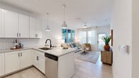 Located at 1900 Elevate Ave in Navarre, FL, this single-floor residence offers a clean, modern aesthetic with a coastal-inspired design. The open-concept layout seamlessly connects the kitchen and living area, creating a bright and airy atmosphere enhanced by light wood-style flooring and crisp white walls throughout.

The kitchen is a standout feature, outfitted with white shaker-style cabinetry, black hardware accents, a classic white subway tile backsplash, and light quartz countertops. Stainless steel appliances — including a range, microwave, refrigerator, and dishwasher — provide full functionality. A central island with seating offers an ideal spot for casual dining or entertaining.

The living area is bathed in natural light from large windows and a glass door that opens to an outdoor balcony. A ceiling fan adds comfort, while the open flow from the kitchen creates an inviting, social space.

The bedroom features soft carpet flooring, a soothing sage green accent wall, natural light from generous windows, and a ceiling fan. The bathroom is finished with clean white tile, a glass-enclosed shower, and light wood-style flooring, maintaining the home's cohesive, serene palette.

Throughout, the design reflects a relaxed Florida coastal lifestyle with thoughtful finishes and a bright, welcoming ambiance.
