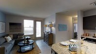 This single-floor residence offers an open-concept design that seamlessly connects the dining and living areas, creating a cohesive and functional layout. The dining area features a granite countertop bar, providing both a casual dining surface and a natural divider between the kitchen and the main living space. Neutral gray walls and light hardwood-style flooring run throughout, lending a clean, contemporary feel to the interior.

The living area is bright and welcoming, benefiting from natural light that filters through large windows fitted with blinds. French-style doors open directly to a private outdoor space, enhancing the sense of openness and allowing for easy indoor-outdoor flow. A warm area rug anchors the seating arrangement, adding texture and comfort to the room.

A decorative console cabinet with twin table lamps serves as an elegant focal point in the transitional space between the living room and the rest of the unit, complemented by wall art that adds a pop of color. Additional rooms are accessible through a hallway visible in the background, suggesting a well-organized and thoughtfully designed floor plan throughout.