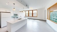 Located on the third floor at 6 Delprete Ave in Rockland, MA, this one-bedroom, one-bathroom unit offers a well-designed, open-concept layout that maximizes its comfortable footprint. The living and kitchen area flow seamlessly together, anchored by a sleek kitchen island with white quartz countertops, white cabinetry, and a soft aqua glass tile backsplash. Stylish pendant lighting hangs above the island, complementing the recessed lighting throughout. Large, wood-trimmed windows flood the space with natural light and offer views of the surrounding neighborhood, enhancing the bright, airy atmosphere created by the crisp white walls and smooth light-toned flooring. The spacious bedroom is accessible through warm-toned solid wood doors and features a large double closet, providing generous storage. A full-wall mirror adds depth to the room, while additional windows continue the theme of natural light. The bathroom is clean and functional, featuring a white vanity with quartz countertop, a wood-framed mirror, and an accessible walk-in shower with grab bars. Warm wood door and trim details throughout the unit add a classic touch that contrasts beautifully with the modern, minimalist finishes.