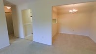 This single-floor residential space offers a clean, functional layout with a bright and neutral aesthetic throughout. The interior is finished with wall-to-wall carpeting that runs seamlessly across the main living areas, lending a warm and comfortable feel underfoot.

The living area flows openly into an adjacent dining space, which is accented by an elegant chandelier that adds a classic touch to the room. A built-in wall niche in the dining area provides a charming architectural detail, ideal for displaying décor or small accessories. The soft, neutral wall tones throughout contribute to a light and airy atmosphere.

A notable feature of this space is the generously sized walk-in closet, visible from the main living area, which is equipped with built-in shelving and hanging rods — offering substantial and well-organized storage. The closet's tiled flooring contrasts with the carpeted main areas, providing a practical surface for a utility or dressing space.

Overall, the space presents a straightforward and versatile interior with modest yet thoughtful details, including architectural niches and dedicated lighting. The open sightlines between rooms enhance the sense of spaciousness, making this a practical and livable environment well-suited for everyday comfort.