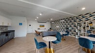 This thoughtfully designed single-floor community space makes a strong visual impression through its signature bold geometric accent walls, featuring a striking blue, beige, and charcoal triangular pattern that spans throughout the room and ties the entire space together cohesively. Light wood-look flooring runs seamlessly underfoot, complementing the clean white ceilings outfitted with recessed lighting and a ceiling-mounted projector for presentations or entertainment.

The lounge is organized into several distinct functional zones. A comfortable seating area hosts two gray sofas accented with patterned throw pillows, arranged around a solid wood coffee table — perfect for casual gatherings. Nearby, teal velvet accent chairs surround marble-topped bistro tables with gold bases, adding a touch of elegance. A dedicated dining and work zone features high-top tables and bar-height seating positioned along large black-framed windows that invite generous natural light and exterior views.

A well-appointed kitchenette includes two-tone cabinetry — white upper cabinets and slate-blue lower cabinets with gold hardware — a dark granite countertop, a farmhouse sink, and a mosaic tile backsplash. A built-in white shelving unit provides display and storage space. The welcoming entry features a keypad-access door, a decorative round mirror, and geometric-patterned floor mat, setting the tone for the stylish interior within.
