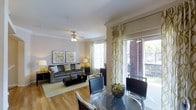 This single-floor residence offers a seamlessly connected living and dining area designed for both comfort and functionality. The open-concept layout is anchored by warm hardwood flooring that flows throughout the main living spaces, creating a cohesive and inviting atmosphere.

The living area features a ceiling fan, elegant crown molding, and large windows dressed with decorative patterned curtains that allow abundant natural light to fill the room. The space comfortably accommodates a full seating arrangement, complete with a sofa, coffee table, and coordinating accent pieces.

Adjacent to the living area, the dining space is defined by a sleek glass-top dining table surrounded by contemporary black chairs, offering a stylish spot for everyday meals or entertaining. A decorative centerpiece adds a refined touch to the table setting.

A standout feature of this residence is the sliding glass door that opens directly onto a private balcony, providing outdoor views of lush greenery and a brick exterior backdrop. The patio offers an ideal extension of the living space for relaxation.

Throughout the home, the neutral color palette, warm tones, and thoughtful design details — including crown molding and coordinated décor accents in yellow and green — contribute to a polished and welcoming ambiance.