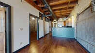 Located at 179 W St Paul Ave in Milwaukee's downtown corridor, this single-floor unit showcases a compelling blend of historic industrial architecture and thoughtfully updated interiors. The open living area immediately impresses with exposed heavy timber ceiling beams, visible ductwork, and rich hardwood flooring that runs throughout, creating a warm yet distinctly urban aesthetic.

The kitchen is a standout feature, anchored by a teal beadboard island that provides both workspace and visual contrast. Natural wood cabinetry reaches toward the ceiling, complemented by a blue tile backsplash, dark granite countertops, and a full suite of stainless steel appliances including a gas range, microwave, and side-by-side refrigerator.

The main living space features an original oversized industrial sliding door set against exposed cream-colored brick, adding authentic character. A separate room offers access to a private balcony through a glass-paned door, flooding the space with natural light and urban views.

Bedrooms feature original brick walls, tall windows with city views, exposed timber columns, and clerestory-style interior windows that borrow light between spaces. The bathroom is finished with brick-tile walls, a dark-toned vanity with black countertop, and a combination tub and shower, maintaining the industrial tone throughout.