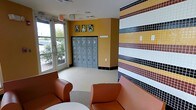 This single-floor facility offers a thoughtfully designed interior that balances functionality with a warm, inviting atmosphere. The main lobby area immediately draws attention with its striking mosaic tile accent wall, featuring a bold horizontal pattern of gold, burgundy, black, white, and gray tiles that adds a distinctive visual character to the space. Warm amber-toned walls complement the vibrant tilework, creating a cohesive and energetic color palette throughout.

The seating area is furnished with comfortable burnt-orange leather armchairs arranged around a small side table, providing a welcoming spot for visitors. Natural light pours in through large glass-paneled entry doors and surrounding windows, offering views of the surrounding greenery outside and enhancing the open, airy feel of the space.

A bank of gray metal lockers lines the back wall, providing practical secure storage for guests. Above the lockers, a series of framed photographs featuring dogs decorates the mustard-yellow wall, reinforcing the pet-friendly nature of the facility. The flooring transitions between tile and carpet, clearly delineating different functional zones within the space. Overall, the facility presents a clean, organized, and cheerful environment well-suited to its purpose.