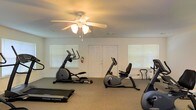 This single-floor fitness space offers a clean and functional environment designed to support a range of cardiovascular workouts. The room features neutral-toned walls and carpeted flooring, creating a comfortable atmosphere for exercise. A ceiling fan with an integrated light fixture is centrally mounted, providing both airflow and overhead illumination to keep the space cool and well-lit during workouts. Multiple windows fitted with white blinds allow natural light to filter in, contributing to an open and airy feel. The fitness area is outfitted with several cardio machines, including an elliptical trainer, a recumbent stationary bike, a treadmill, and additional exercise equipment, offering users a variety of workout options. A set of double doors provides convenient access, and a fire extinguisher is visibly mounted near the entrance, reflecting attention to safety. The layout allows for ample space between machines, supporting comfortable use and ease of movement throughout the room. Overall, this fitness space strikes a balance between practicality and accessibility, making it a straightforward and inviting area for regular exercise.