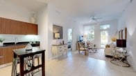 This single-story residence is characterized by its airy, open-concept design and elevated ceilings that lend a spacious, expansive feel throughout. Large-format light tile flooring flows seamlessly across the main living areas, reinforcing the sense of openness and continuity.

The living room is generously proportioned, bathed in natural light streaming through floor-to-ceiling windows and a glass door that frames lush outdoor greenery. A ceiling fan overhead adds a casual, comfortable touch to the bright, neutral-toned space. The adjoining dining area accommodates a full dining set beneath a pendant light fixture, positioned to take advantage of the open sightlines into the living room.

The kitchen is a standout feature, outfitted with rich wood-grain cabinetry extending to the ceiling, dark granite countertops, under-cabinet lighting, a glass tile backsplash, and a full suite of stainless steel appliances. A compact bistro-style table and stools serve as a casual dining option within the kitchen itself.

Both bedrooms are carpeted and feature large windows with outdoor views, offering a calm, restful atmosphere. Each bathroom is finished with matching wood-grain vanities, dark granite countertops, framed mirrors, and tiled tub-shower combinations, maintaining a consistent, refined aesthetic throughout the residence.