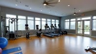 This fitness center is a thoughtfully designed exercise space built to accommodate a variety of cardio-focused workouts. The room features warm hardwood flooring that extends throughout, providing a clean and durable surface well-suited for both machine-based and open-floor exercises. A row of large windows lines the walls, flooding the space with abundant natural light and offering views of the surrounding greenery, creating an inviting and energizing atmosphere. The walls are finished in a cool grey tone that complements the modern aesthetic of the room. The ceiling is fitted with recessed lighting alongside ceiling fans, ensuring the space remains well-lit and well-ventilated during use. The equipment lineup includes multiple treadmills, stationary upright and recumbent bikes, and additional cardio machines, each equipped with individual display screens for an enhanced workout experience. The open layout provides ample floor space between machines, allowing for comfortable movement and accessibility. Overall, this fitness center offers a bright, modern, and functional environment designed to support an active and healthy lifestyle.