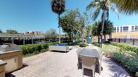 This inviting single-level outdoor space exudes a relaxed, tropical ambiance characteristic of South Florida living. The expansive courtyard is finished with decorative interlocking paver stones that lend a refined, clean aesthetic underfoot. The space is thoughtfully arranged with distinct seating zones: a cushioned wicker lounge set with soft blue upholstery provides a comfortable area for casual relaxation, while a sleek rectangular dining table with wicker chairs offers a dedicated spot for outdoor dining and gathering. Mature palm trees and a variety of lush tropical greenery frame the perimeter, providing natural shade and privacy while contributing to the vibrant, resort-like atmosphere. Well-maintained hedgerows and manicured shrubs define the boundaries of the space, complemented by classic black iron fencing and traditional-style lamp posts that add a touch of charm. Glimpses of neighboring architecture and a clear blue sky above enhance the open, airy feel. The overall environment is bright, welcoming, and ideally suited for outdoor entertaining or leisurely enjoyment in a warm, sunny climate.