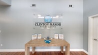 The Clinton Manor Apartments leasing office is a thoughtfully designed, single-floor space that serves as the welcoming hub for the community. Upon entry, visitors are greeted by a prominent branded glass sign and a natural wood display table showcasing floor plans and community information, setting a professional yet inviting tone.

The main area is anchored by a striking floor-to-ceiling brick fireplace, which adds warmth and character to the space. Exposed ceiling beams and pendant lighting complement the cozy seating arrangement of rich brown leather chairs gathered around a coffee table, creating a comfortable lounge area for prospective residents. Rich hardwood flooring flows throughout, tying the space together cohesively.

The office workspace features a dedicated leasing desk positioned against a wall of large French doors and sliding glass panels, flooding the area with natural light and offering views of the surrounding greenery. The soft blue-grey walls throughout contribute to a calm, professional ambiance.

A well-appointed kitchenette is tucked within the space, outfitted with warm oak cabinetry, dark granite countertops, and a stainless steel sink — a practical amenity for staff and visitors alike. A small drop-leaf table and bar stools provide an additional informal meeting area. Together, these elements create a functional, attractive, and welcoming leasing environment.
