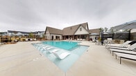 Located at 37 Foxgrove Dr in Dawsonville, Georgia, this single-story community amenity facility showcases a modern farmhouse architectural style, characterized by board-and-batten siding, a pitched shingle roof, and dark-framed windows that lend a refined, contemporary aesthetic. At the heart of the space is a generously sized resort-style swimming pool, featuring built-in in-water lounge chairs for relaxed sunbathing and steps for easy entry. The expansive pool deck is lined with chaise lounges and shaded by umbrellas, providing ample space for outdoor recreation and leisure. Flanking the pool are two covered outdoor living areas supported by rustic wood columns and finished with shiplap ceilings and ceiling fans for year-round comfort. One covered porch features a striking full-height stacked stone fireplace with a mounted flat-screen television, surrounded by sophisticated neutral-toned seating, creating an inviting gathering space. The second covered patio area includes a walk-up bar window with counter seating, woven lounge chairs, and a fire pit, ideal for casual socializing. The entire outdoor area is enclosed by black iron fencing, with mature trees providing a natural backdrop and views of the surrounding residential neighborhood.