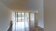This single-floor space offers an open-concept layout highlighted by rich hardwood flooring that reflects natural light throughout. The living area is anchored by expansive floor-to-ceiling sliding glass doors fitted with vertical blinds, offering direct access to a private balcony overlooking a verdant, tree-lined outdoor setting. A second set of vertical blinds covers an additional window, further enhancing the sense of openness and light. The neutral-toned walls and textured ceiling complement the warm tones of the flooring, creating a cohesive and inviting atmosphere. Flush-mount ceiling lighting provides functional illumination, while the generous proportions of the main living space allow for flexible furniture arrangements. The balcony, accessible through the sliding doors, extends the living area outdoors and provides pleasant views of the surrounding greenery. Overall, the space presents a clean, well-lit environment with an emphasis on natural light, durable finishes, and a seamless connection between interior and exterior living.