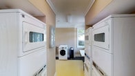 This single-floor laundry room is designed for shared use, featuring a practical and well-organized layout. The space is lined on both sides with stacked washer and dryer units, maximizing capacity within the room's footprint. Additional freestanding front-load washers are positioned toward the back of the room, providing further flexibility for users.

The room is finished with warm beige walls and crown molding near the ceiling, lending a polished, clean aesthetic uncommon in utility spaces. The light yellow flooring adds a bright, welcoming tone underfoot. A coffered ceiling detail with recessed paneling provides a subtle architectural element that elevates the overall feel of the room.

Natural light enters through multiple windows along the back wall, keeping the space feeling open and well-lit. A small folding or utility table near the back wall offers a convenient surface for sorting and folding laundry. A waste bin is also present for added convenience. The windows overlook greenery outside, contributing to a pleasant and functional environment within this dedicated laundry facility.