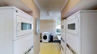 This single-floor laundry room is designed for shared use, featuring a practical and well-organized layout. The space is lined on both sides with stacked washer and dryer units, maximizing capacity within the room's footprint. Additional freestanding front-load washers are positioned toward the back of the room, providing further flexibility for users.

The room is finished with warm beige walls and crown molding near the ceiling, lending a polished, clean aesthetic uncommon in utility spaces. The light yellow flooring adds a bright, welcoming tone underfoot. A coffered ceiling detail with recessed paneling provides a subtle architectural element that elevates the overall feel of the room.

Natural light enters through multiple windows along the back wall, keeping the space feeling open and well-lit. A small folding or utility table near the back wall offers a convenient surface for sorting and folding laundry. A waste bin is also present for added convenience. The windows overlook greenery outside, contributing to a pleasant and functional environment within this dedicated laundry facility.