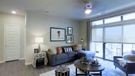 This single-floor space showcases a well-appointed living area designed with contemporary sensibilities. The room features hardwood-style flooring that adds warmth and visual continuity throughout. A large window dressed with patterned curtains and a roller shade floods the space with natural light, while offering views to an adjoining balcony accessible through a sliding glass door. The ceiling is fitted with a ceiling fan, providing both functional airflow and a subtle decorative element. The living area is centered around a spacious sectional sofa in a neutral gray tone, accented with an eclectic mix of throw pillows in rust, striped, and solid patterns. A round, dark-wood coffee table anchors the seating arrangement, complemented by a textured pouf that adds an informal, layered aesthetic. Wall art is thoughtfully arranged in a gallery-style grouping, adding personality to the neutral-toned walls. A floor lamp and side table with decorative accessories complete the setting, contributing to a cozy yet curated ambiance. Overall, the space balances modern design with lived-in comfort.