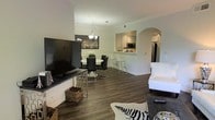 This well-appointed single-floor residence offers a seamless open-concept layout that connects the living, dining, and kitchen areas into one cohesive, flowing space. Rich, dark hardwood floors run throughout, grounding the interior with warmth and sophistication. The living area is anchored by a large flat-screen television mounted on a sleek media console, complemented by a cozy white sofa and a bold zebra-print area rug that adds a contemporary flair. The dining area features a round white table paired with dark upholstered chairs, positioned beneath an elegant chandelier that casts a warm glow. Crown molding runs along the ceilings, adding a refined architectural touch throughout the space. A distinctive arched passageway leads into the kitchen, which is outfitted with white cabinetry, stainless steel appliances — including a microwave and refrigerator — and a raised breakfast bar with modern bar stools, ideal for casual dining. The kitchen's pass-through design keeps the space connected and airy. Overall, the residence strikes a sophisticated balance between modern design and comfortable living, offering a polished and inviting atmosphere throughout its open, well-organized layout.