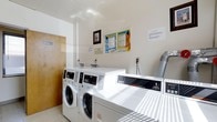 This single-story laundry facility provides a functional and well-organized space designed to accommodate shared laundering needs. The room features commercial-grade front-loading washing machines and dryers positioned along the walls, offering efficient and reliable performance. The interior is finished with crisp white walls and light-toned flooring, contributing to a clean and hygienic environment. Instructional signage and usage guidelines are clearly posted on the walls, ensuring residents can operate the machines with ease and confidence. A ventilation duct is visibly integrated into the layout, supporting proper airflow for the drying equipment. The space is illuminated by overhead lighting that brightens the room evenly, maintaining visibility throughout. A wood-paneled door provides access to the facility, and the overall layout is straightforward and efficiently arranged to allow multiple users to navigate comfortably. The no-frills, utilitarian design prioritizes function and cleanliness, making this laundry room a practical and essential amenity.