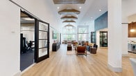 Located at 14885 Memorial Dr in Houston, TX, this thoughtfully designed single-floor amenity space offers a seamless blend of comfort, style, and functionality across its generously sized layout. The main living room is an immediate showstopper, boasting soaring double-height ceilings and floor-to-ceiling gridded windows that flood the room with natural light and frame lush outdoor greenery. Light wood flooring, a sculptural sputnik chandelier, and contemporary furnishings give the space a sophisticated yet inviting atmosphere. An adjacent lounge area features curated abstract artwork, high ceilings with subtle beam accents, and warm hardwood floors, creating an elegant retreat for socializing or relaxing. The kitchen and living room area is a standout, showcasing sage green cabinetry paired with rich wood-slat upper panels, white stone countertops, globe pendant lighting, and a herringbone-patterned hardwood floor — all combining to deliver a warm, modern aesthetic with premium finishes. The expansive exercise room is comprehensively outfitted with cardio machines including treadmills and ellipticals, a full suite of strength equipment, free weights, and large arched mirrors, all set on durable sport flooring with ample natural light from surrounding windows. A connecting hallway provides smooth circulation throughout the space, tying each area together with clean, modern design continuity.