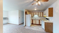 This single-floor residence offers a practical and functional layout centered around an open-concept design that connects the kitchen and living area seamlessly. The kitchen is well-appointed with cream-colored cabinetry accented by warm wood-toned trim, providing ample storage space across both upper and lower cabinets. White appliances, including a full-size refrigerator, electric range, and dishwasher, are neatly integrated into an L-shaped counter configuration. An under-cabinet light above the sink adds task lighting, while a flush-mount ceiling fixture provides general illumination. The transition from the tile flooring in the kitchen to the soft, neutral carpeting of the living area creates a clear and comfortable division between the two spaces. The living area benefits from a ceiling fan with a light kit, contributing to both comfort and ambient lighting. The walls are finished in a clean, light tone that enhances the overall brightness of the interior. Straightforward baseboards and trim details complete the look, giving the space a tidy, well-maintained character. The open flow between the kitchen and living area makes this a highly livable and adaptable single-floor home.