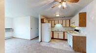This single-floor residence offers a practical and functional layout centered around an open-concept design that connects the kitchen and living area seamlessly. The kitchen is well-appointed with cream-colored cabinetry accented by warm wood-toned trim, providing ample storage space across both upper and lower cabinets. White appliances, including a full-size refrigerator, electric range, and dishwasher, are neatly integrated into an L-shaped counter configuration. An under-cabinet light above the sink adds task lighting, while a flush-mount ceiling fixture provides general illumination. The transition from the tile flooring in the kitchen to the soft, neutral carpeting of the living area creates a clear and comfortable division between the two spaces. The living area benefits from a ceiling fan with a light kit, contributing to both comfort and ambient lighting. The walls are finished in a clean, light tone that enhances the overall brightness of the interior. Straightforward baseboards and trim details complete the look, giving the space a tidy, well-maintained character. The open flow between the kitchen and living area makes this a highly livable and adaptable single-floor home.