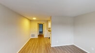 This single-floor unit offers a clean, functional layout designed for comfortable everyday living. The open living and dining area is finished with warm, light-toned hardwood-style flooring that extends through much of the space, creating a cohesive and easy-to-maintain surface underfoot. Neutral-toned walls throughout provide a versatile backdrop suited to a variety of furnishing styles.

The kitchen is efficiently arranged with white cabinetry offering ample storage, granite-style countertops, a double stainless steel sink, and black appliances including an electric range and refrigerator. Recessed lighting adds a bright, functional atmosphere to the cooking area, which opens toward the main living space.

A hallway connects the living areas to the additional rooms, one of which features plush gray carpeting and a window that brings in natural light along with views of greenery outside. The entry area includes a solid paneled door with a window that allows natural light to filter in, enhancing the brightness of the space.

Ceiling-mounted HVAC vents and floor-level returns are present throughout, indicating a centralized climate control system. The overall layout, as seen in the dollhouse view, is compact yet well-organized, with clearly defined zones for living, cooking, and private use.