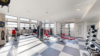 Located at 5727 North 7th Street in Phoenix, AZ, this fitness center offers a generously sized, single-floor exercise room designed with both functionality and aesthetics in mind. The space features soaring ceilings with floor-to-ceiling windows along multiple walls, flooding the interior with abundant natural light and offering open views of the surrounding Phoenix cityscape.

The exercise room is finished with a striking two-tone checkerboard tile floor in shades of grey, lending the space a clean, modern character. The walls are a crisp white, complemented by exposed concrete block accents that add an industrial touch. Full-length mirrors are strategically placed to enhance the sense of openness and support workout visibility.

Equipment is comprehensive and well-organized throughout the space. Cardio machines from True Fitness and Life Fitness — including treadmills, ellipticals, and stair climbers — are arranged along the windowed walls to take advantage of natural light. A dedicated free weights area features a full dumbbell rack and flat-screen TV mounted to the wall. Additional strength training equipment, including cable machines and multi-functional weight stations with red upholstered benches, rounds out the offering. Recessed lighting supplements natural light throughout, and the space is finished with a clean, professional ambiance well-suited for a focused fitness experience.