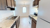 This single-floor space centers around a galley-style kitchen that is both practical and well-appointed. The kitchen features dark brown cabinetry lining both walls, providing ample storage above and below the countertops. The counters display a marbled laminate surface, offering generous prep space on either side of the layout. A stainless steel sink with a single basin is positioned along the left wall, accompanied by a dishwasher tucked neatly beneath the counter. On the right side, a white gas range with multiple burners serves as the primary cooking appliance, complemented by an overhead range hood for ventilation. The light-toned vinyl tile flooring runs throughout, contributing to a clean and cohesive look. Natural light enters through a window at the far end of the kitchen, framed by vertical blinds, creating a bright focal point in the room. A decorative pendant light hangs above this window, adding a subtle design element to the space. The white walls throughout maintain a neutral, open atmosphere, making the darker cabinetry stand out as the defining design feature of this functional kitchen space.